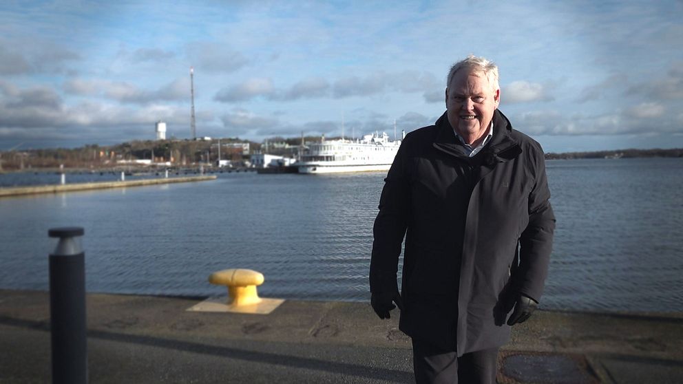 En person i svart jacka går längs en strandpromenad vid en hamn, med båtar som ligger förankrade i bakgrunden. Himlen är delvis molnig och vattnet verkar stilla.