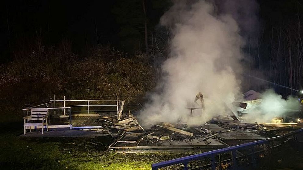 Rök stiger från resterna av en nerbrunnen byggnad i en skogsmiljö under natten. Brända träbjälkar och plåt ligger utspritt på marken, medan rök och dimma skingras i luften. En ljuskälla kastar sken på vraket.