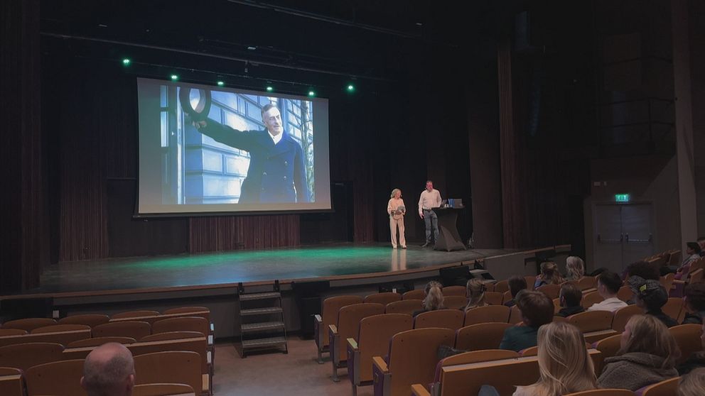 Två personer håller presentation i en salong med åhörare och stor bildskärm i bakgrunden.