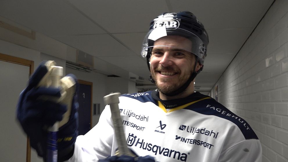 Oscar Engsund i en omklädningshall håller en klubba och en annan utrustning. Han är klädd i en tröja med sponsormärken och en hjälm, vilket antyder att han förbereder sig för en match eller träning. Bakgrunden är neutralt grå med belysning längs korridoren.