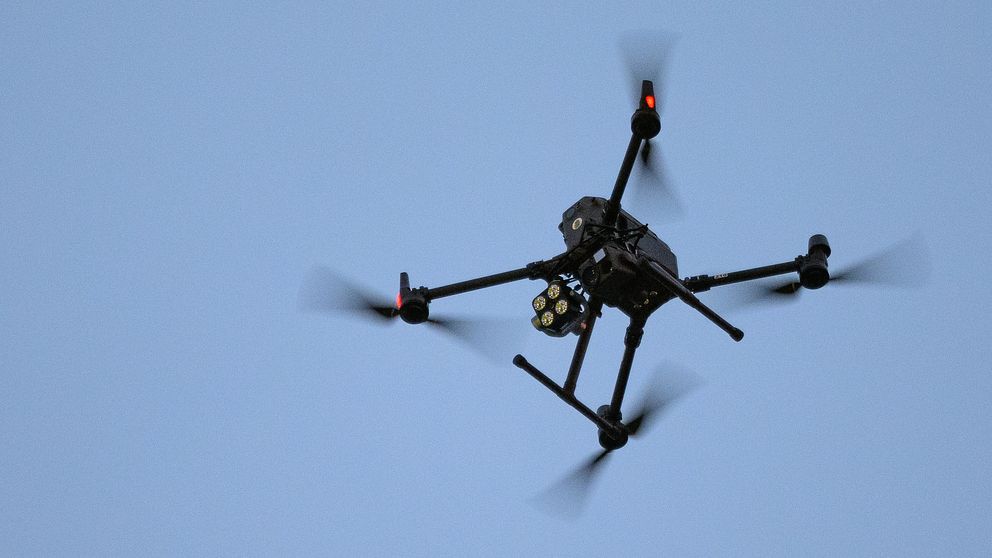 En svart drönare med roterande propellrar sväva i en klar himmel, synlig med tre av sina fyra ben och en kamera under kroppen.