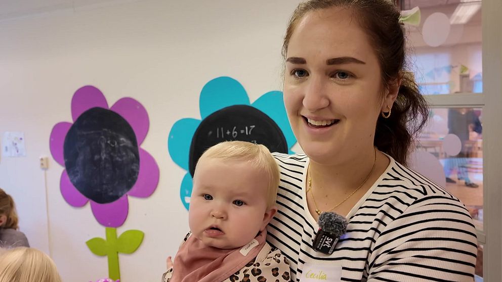 En kvinna håller ett litet barn i en förskolemiljö med färgglada blommor på väggarna. I bakgrunden finns tavlor i svart som kan användas för undervisning. Atmosfären verkar varm och inbjudande, vilket tyder på en lärorik och trygg plats för barn.