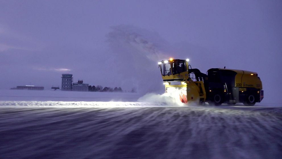 Snöröjare i aktion på en snötäckt flygplats under skymning, med kraftfulla lampor som lyser upp arbetsområdet medan snön blåser bort från banan. I bakgrunden syns en byggnad som kan vara en flygkontroll.