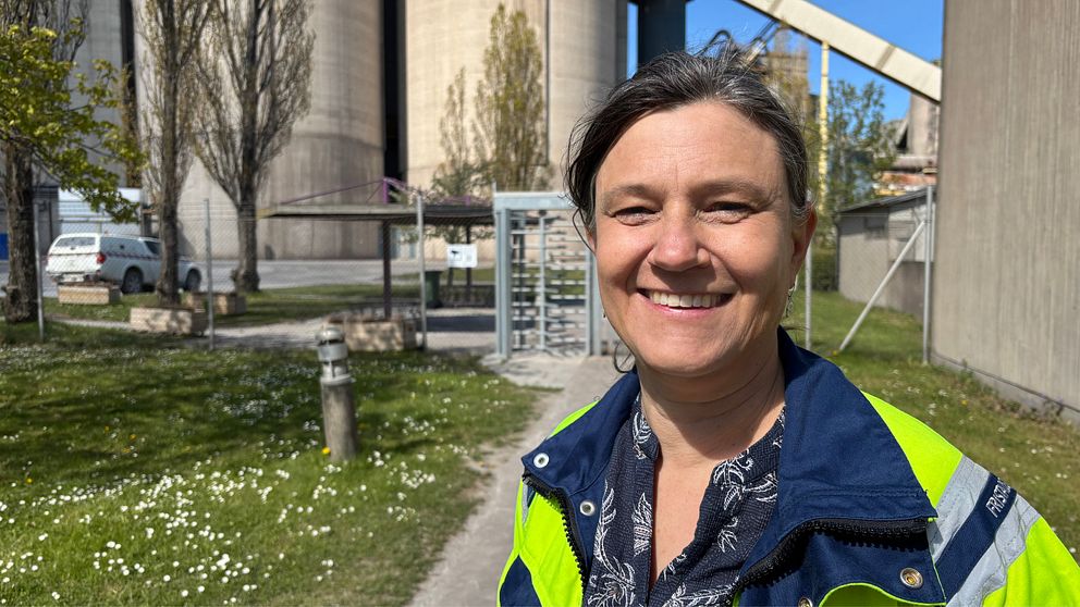 Leende person med reflexjacka utomhus vid industrimiljö med silos och grön gräsmatta.