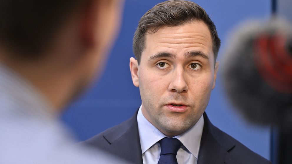 Sveriges biståndsminister Benjamin Dousa intervjuas. Han har en mörkblå kostym och står framför en mikrofon, till synes intervjuad av en annan person vars halva bakhuvud syns i förgrunden. Bakgrunden är enfärgat blå.