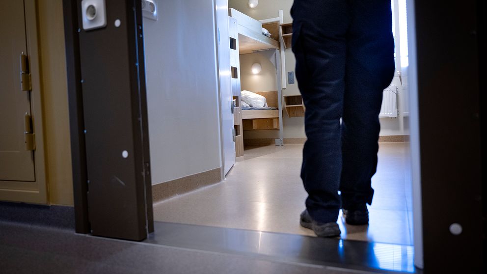 En person i arbetskläder går in i en cell i ett modernt fängelse. Cellen är inredd med två våningssängar och har ett fönster som släpper in lite ljus. Dörren till cellen är delvis öppen, vilket ger en vy av den minimalistiska inredningen.