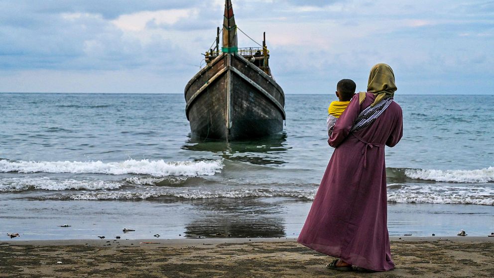 En kvinna i lång klänning med ett barn i famnen står på en strand och ser på en båt.