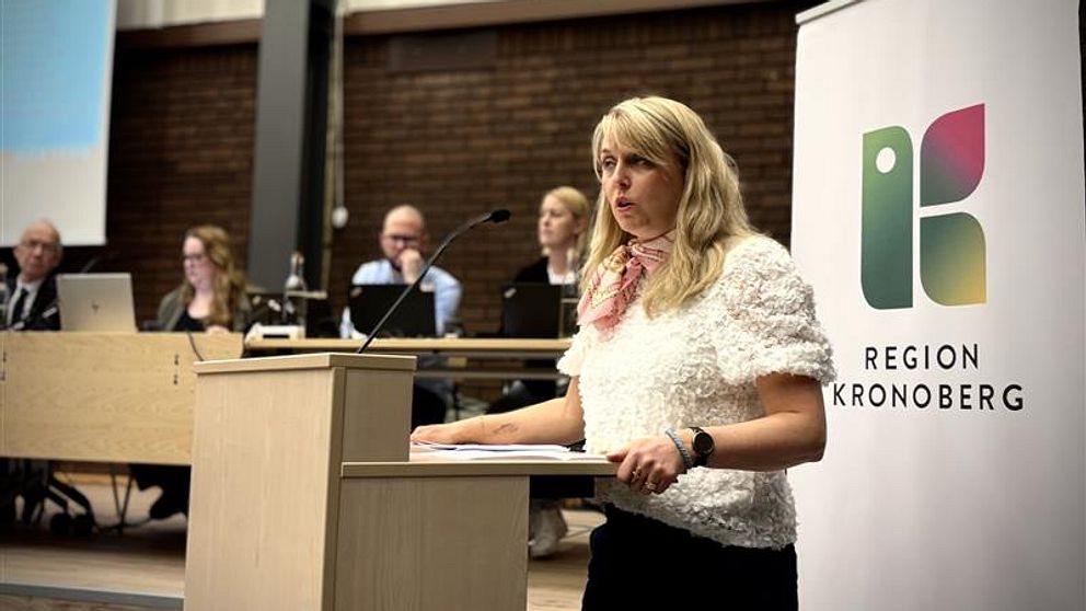 Anna Zelvin (KD) vid podium talar. Region Kronoberg skylt i bakgrunden. Människor sitter vid bord.
