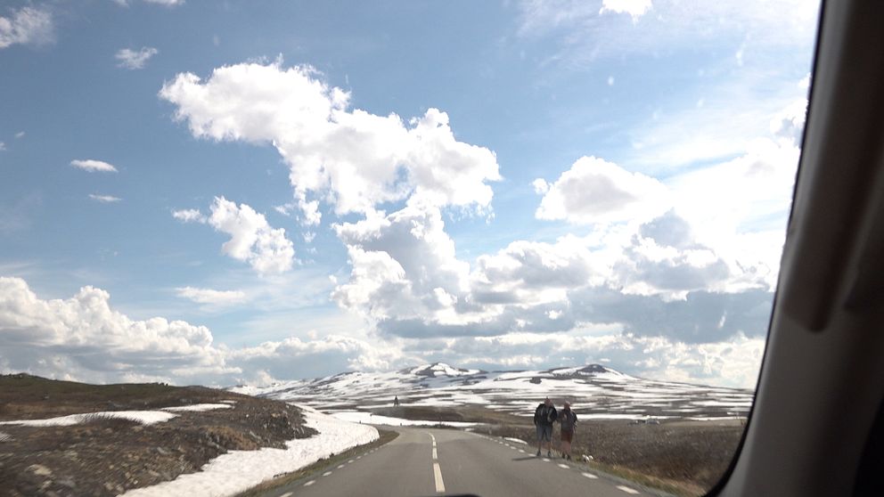 Landsväg genom fjällandskap med snö, två vandrare ses längre fram på vägen.