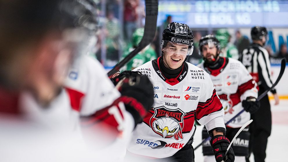 Ishockeyspelare i vita och röda tröjor på isen under en match. Domaren syns bakom.