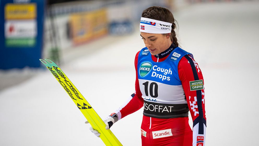 Skidåkare i röd tävlingsdräkt bär skidor på snötäckt arena vid tävling.