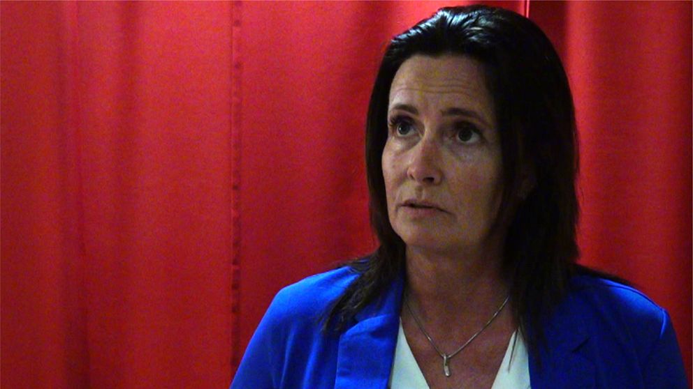 En person i en blå kavaj och vit blus står framför en röd bakgrund och håller i en mikrofon. Situationen tyder på att det handlar om en intervju eller presskonferens.