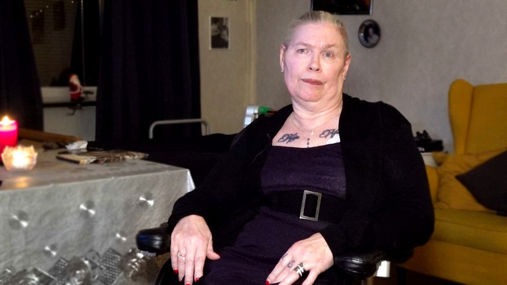 En person sitter avslappnat i en rullstol, klädd i en svart jacka över en lila klänning. I bakgrunden syns en mysig inredning med mörka gardiner och en gul soffa. Bredvid personen finns ett bord med några tända ljus och dekorationer, vilket skapar en varm atmosfär.