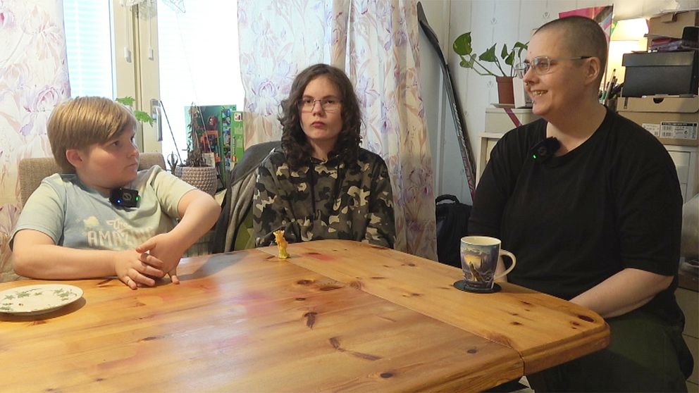 Tre personer, två pojkar och en förälder, sitter vid ett köksbord med muggar och tallrik i hemmiljö.