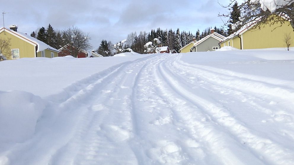 En snöbelagd väg som leder genom ett bostadsområde med gula hus på sidorna, omgiven av orörd snö och träd i bakgrunden.