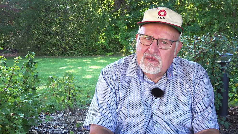 En man, Kent Ossiansson som är ordförande för FUB södra Sörmland, sitter på en bänk i en grönskande trädgård omgiven av buskar och gräs. Han bär en ljusblå skjorta med mönster och en keps. Soligt väder och en lugn atmosfär präglar scenen.