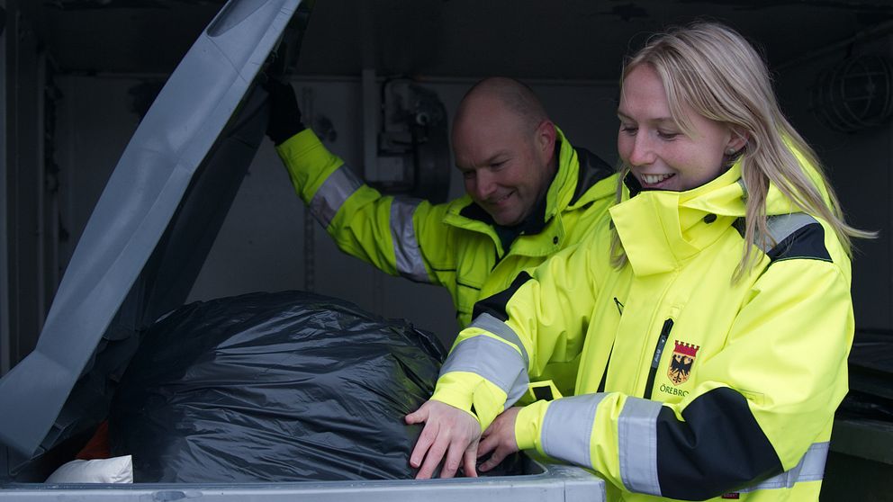 Två personer i neonfärgade arbetsjackor är i färd med att tömma en soptunna, där en stor svart sopsäck syns. Den ena personen sträcker sig in i tunnan för att hantera avfallet.