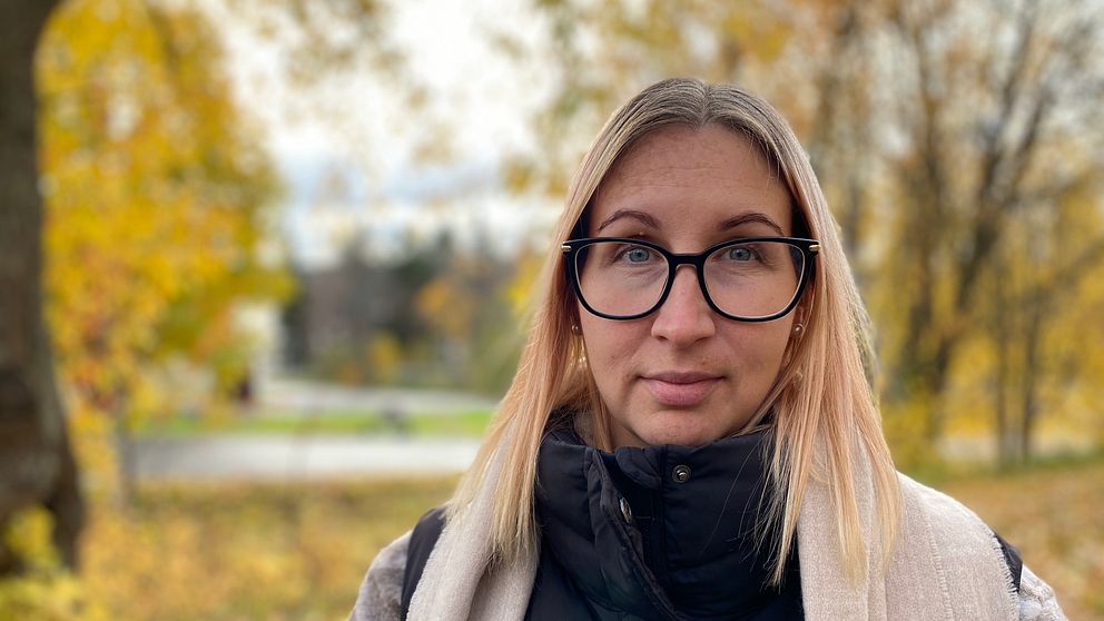 En person står i en skogsmiljö med höstfärger. Bakgrunden visar träd med gula och gröna löv samt en väg som sträcker sig bortom. Personen är klädd i en svart jacka och en ljus sjal, vilket ger ett avslappnat intryck i den naturnära omgivningen.