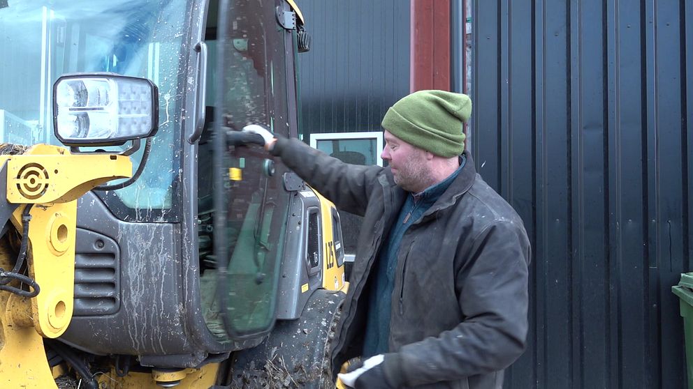 En person står vid en maskin och öppnar dörren. Maskinen har smutsiga ytor, vilket tyder på att den har använts i ett jord- eller byggarbetsprojekt. I bakgrunden syns en vägg av svart plåt, vilket ger en industriell känsla.
