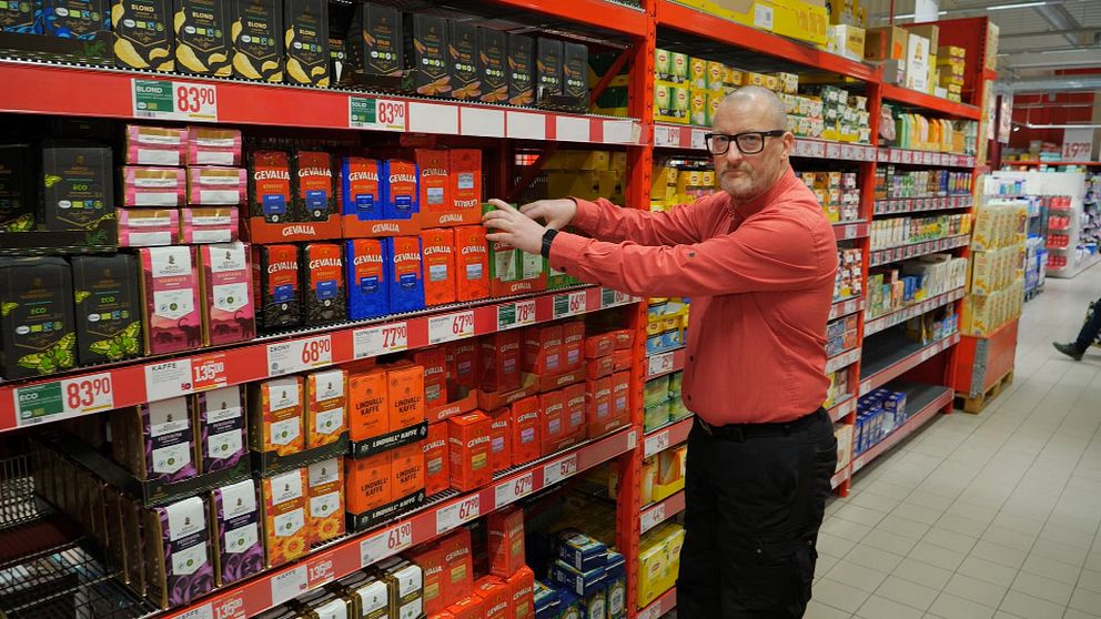 en man i röd skjorta sorterar kaffepaket på en hylla i en butik