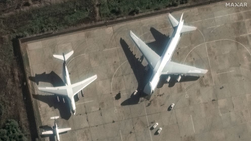 Två flygplan står parkerade på en flygplats. Det mindre flygplanet är placerat till vänster, medan det större flygplanet är till höger. Markytan är synlig i bakgrunden med en del av omgivande vegetation. Små fordon rör sig nära flygplanen.