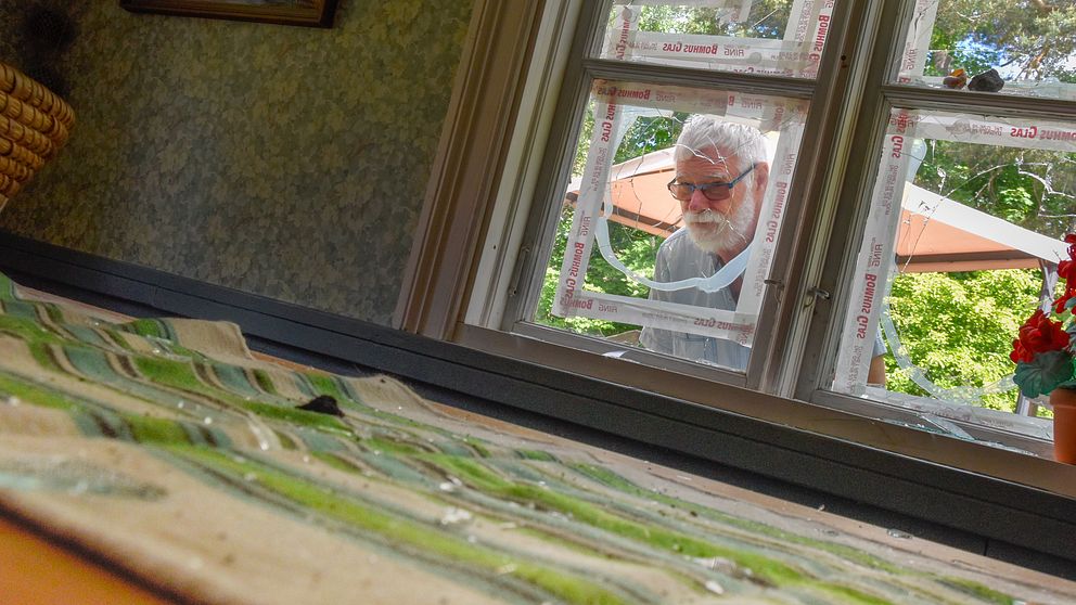 Hembygdsföreningens ordförande Hans Wahlbom tittar in genom ett fönster som krossats av stenar. På ett bord inne i huset ses glassplitter.