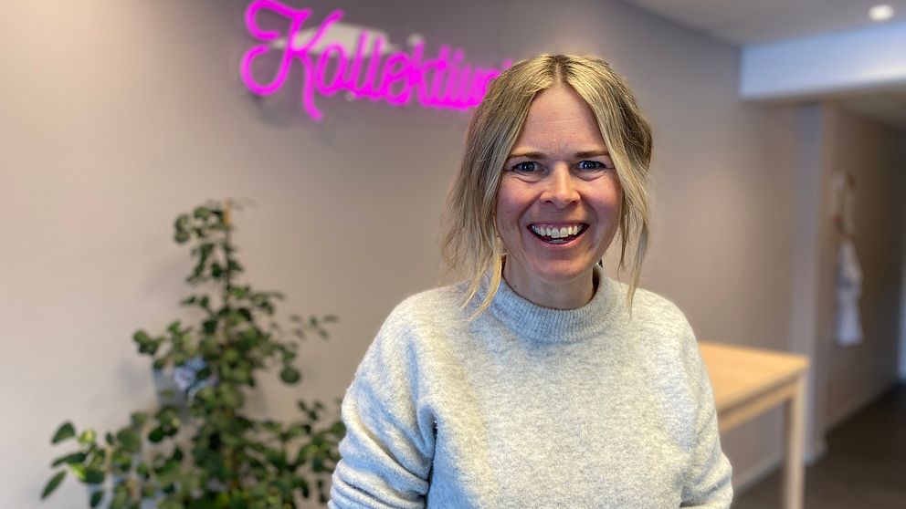 Jenny Ståhl, blond kvinna med uppsatt hår och grå tröja. I bakgrunden en neonskylt med texten ”Kollektivet”