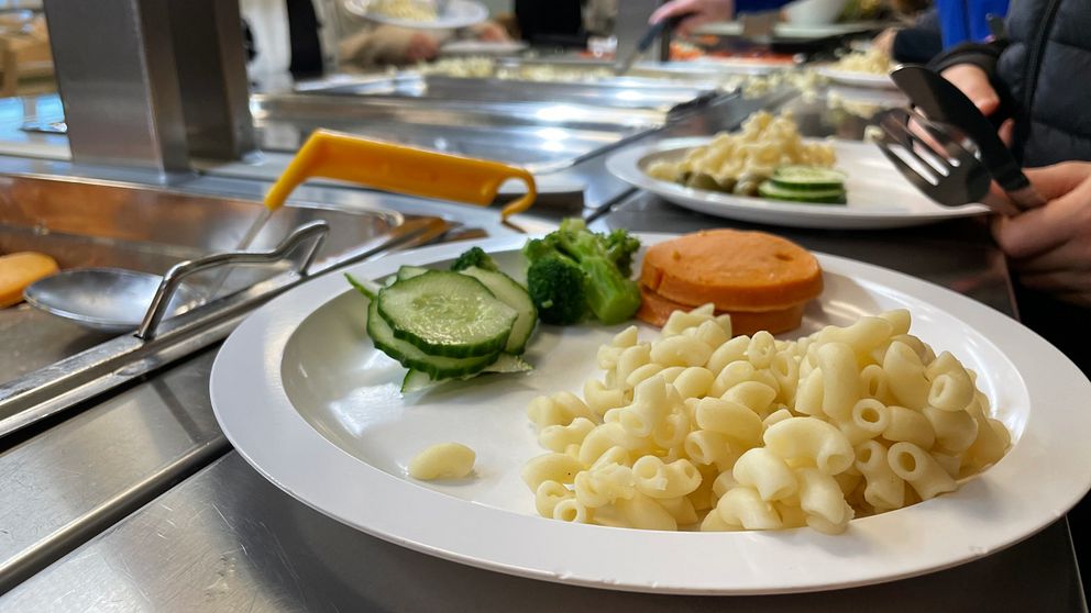 En tallrik med pasta, broccoli, gurkskivor och en bit korv, placerad på en serveringsdisk i en matsal. I bakgrunden syns andra maträtter och personer som serverar sig själva.