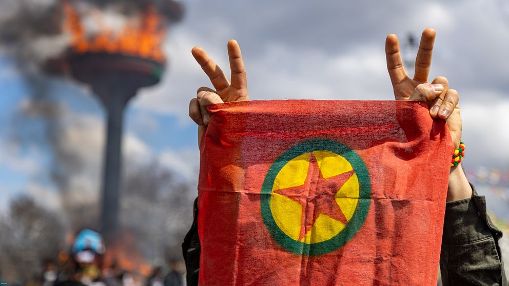 Person håller PKK-flagga och gör segertecken, brinnande fackla i bakgrunden.