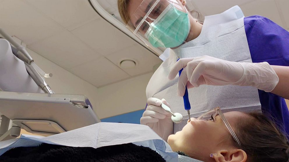 En tandvårdspersonal som arbetar med en patient under en tandbehandling. Tandläkaren håller ett instrument och fokuserar på patientens mun, medan det finns olika verktyg synliga i arbetsområdet. Patienten ligger på en behandlingsstol, och säkerhetsåtgärder som skyddande kläder används.