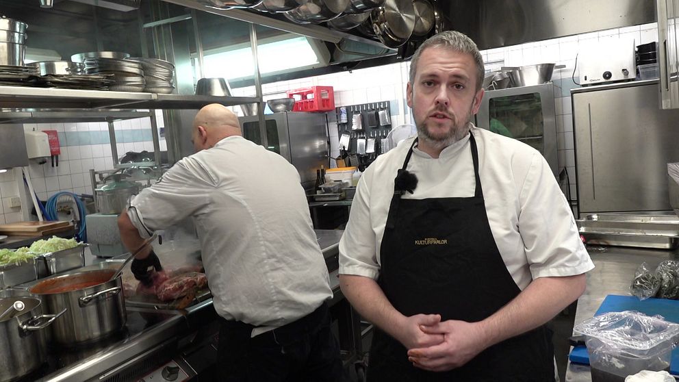 Kökschefen Andreas Eriksson står i köket och tittar mot kameran. I bakgrunden syns en kock steka kött.
