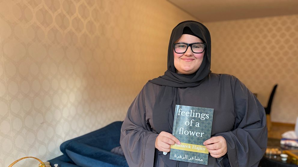 En person i en lång, mörk klädsel håller en bok med titeln ”feelings of a flower”. I bakgrunden syns en blå soffa och inredning som skapar en hemtrevlig atmosfär. Det ligger några böcker och föremål på ett bord i närheten.