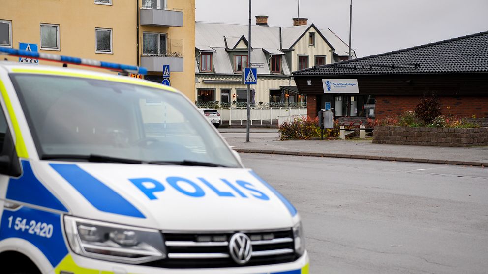 En polisbil i fokus på en gata, med byggnader i bakgrunden, inklusive en butik och flerfamiljshus. Orten verkar vara en mindre stad med en lugn atmosfär.