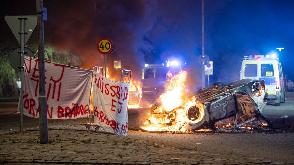Bild från upploppet i Rosengård efter koranbränningarna i Malmö. Till vänster banderoll med texten: ”Nej till koranbränningar”, bredvid en brinnande bil och polisbil.