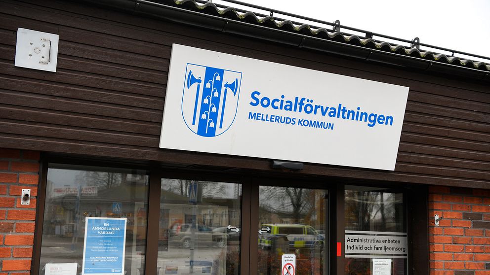 Entrén till Socialförvaltningen i Melleruds kommun, med en skylt som tydligt visar namnet på institutionen. Flera informationsskyltar och dokument är synliga på fönstren. Byggnaden har en tegelvägg och ett platt tak, i en urban miljö.