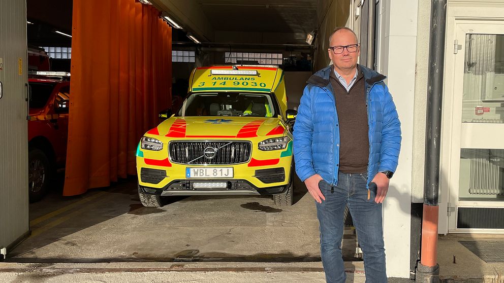 Man med glasögon och blå jacka står framför ett ambulansgarage.