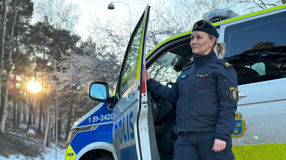 Polis som står vid polisbil tittar ut.