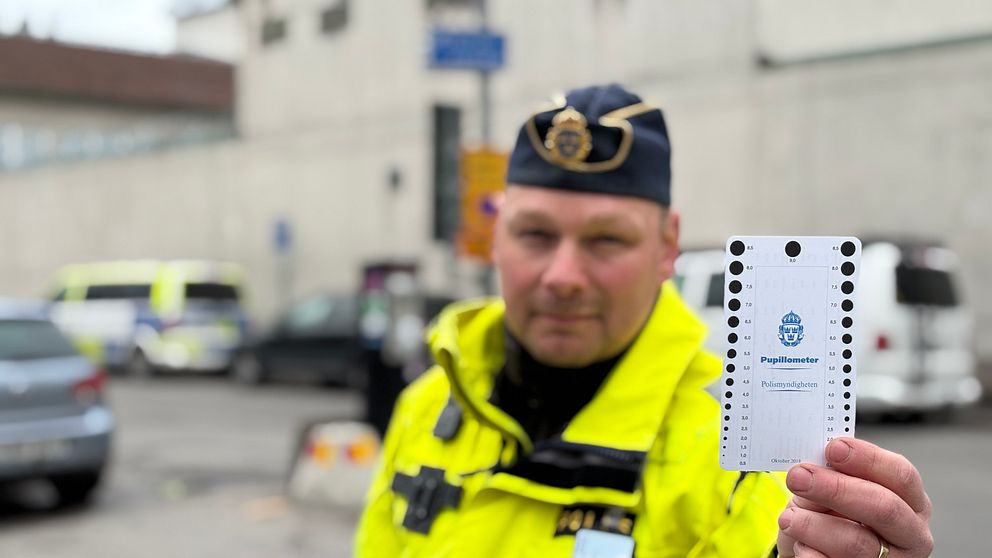 Polis i gul jacka håller upp ett polisdokument på en gata med bilar i bakgrunden.