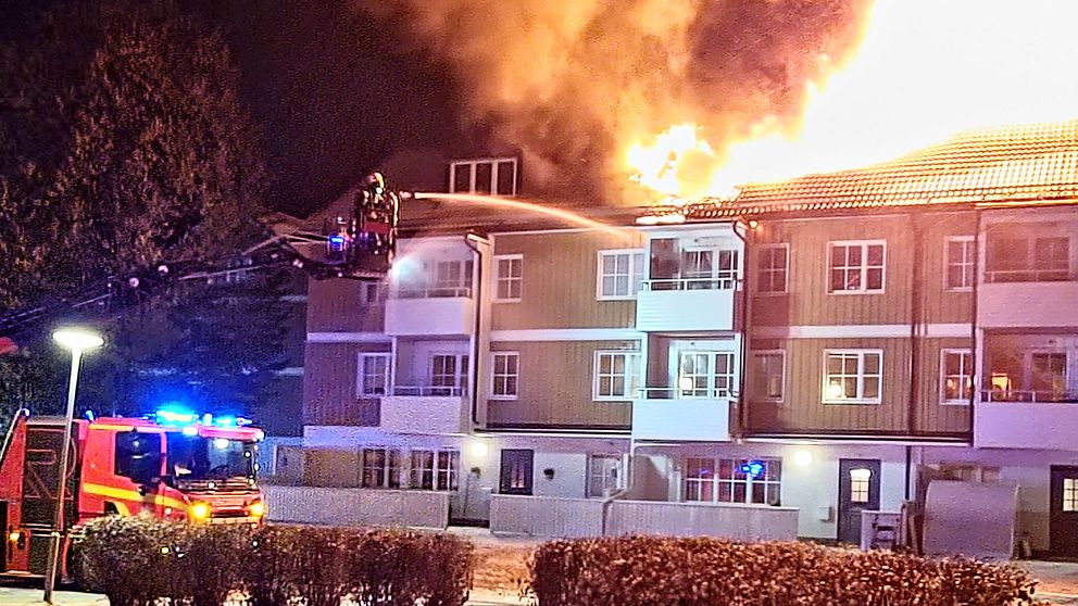 Lågor slår upp från taket på ett två-våningshus och räddningstjänstens sprutar vatten mot en balkong från en stegbil.