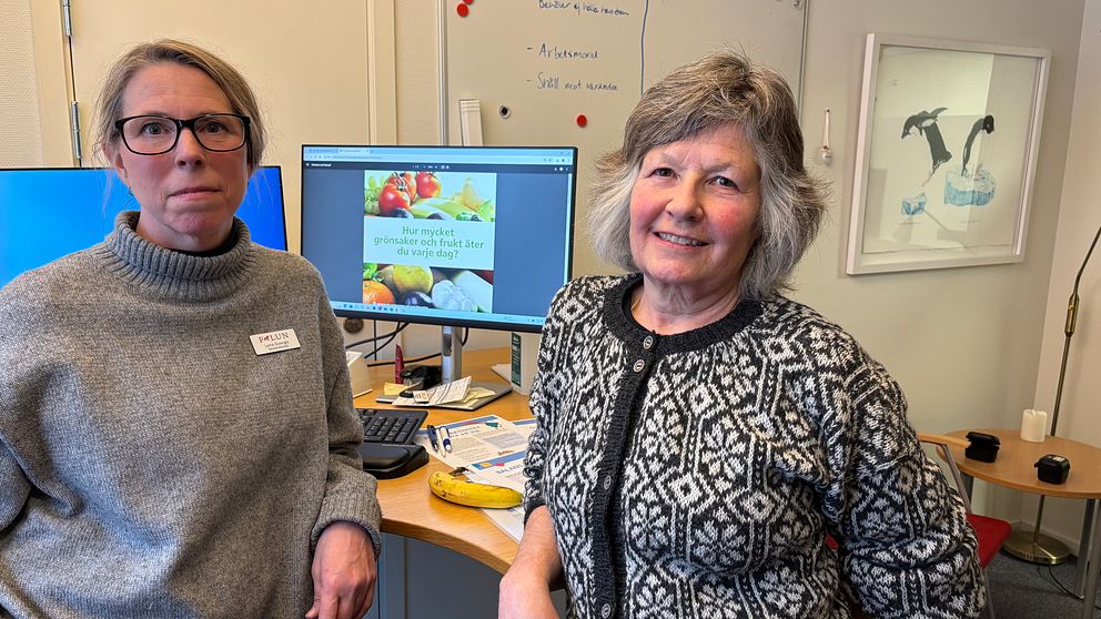 Två skolsköterskor står bredvid ett skrivbord i ett kontor. På en datorskärm syns råd om hälsosamma livsmedel som frukt och grönsaker.