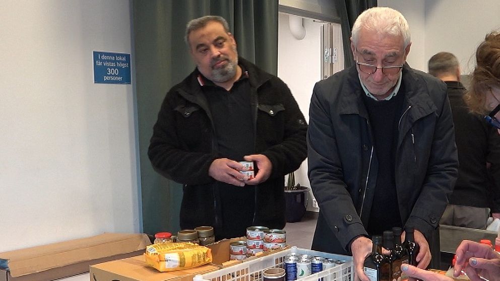 Två män i svarta jackor packar upp matvaror på ett bord. En kvinna med telefon köper en vara.