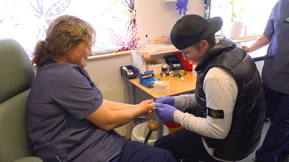 En ung pojke som går i åttonde klass, med bakvänd keps och svart dunväst, provar på att ta ett blodprov i fingret på en sjuksköterska