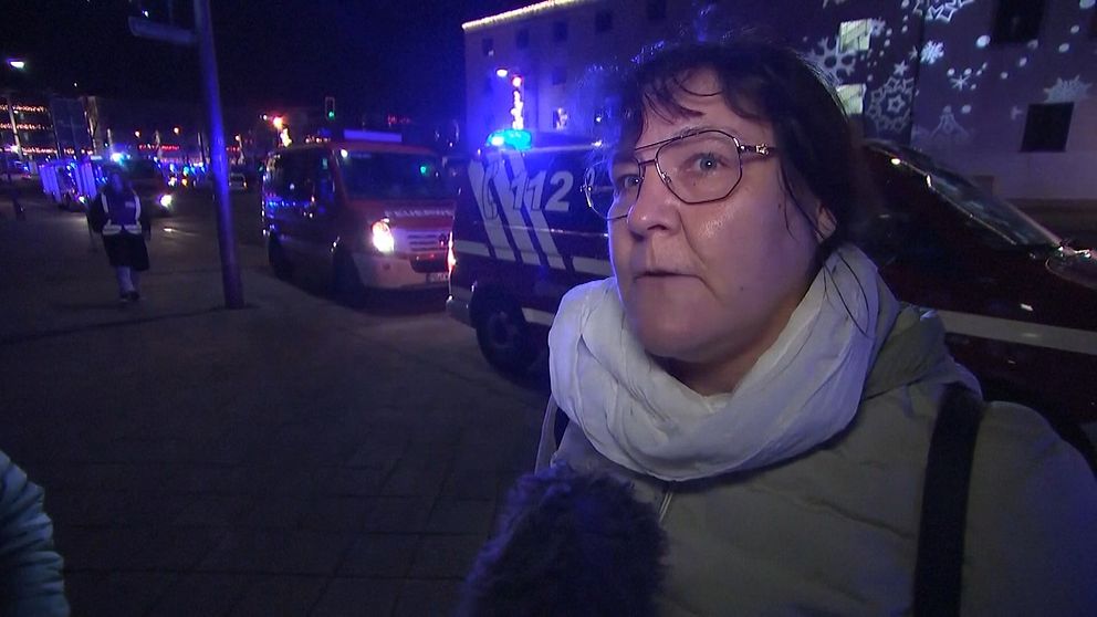 En person står på gatan med en vit scarf, medan räddningstjänstens fordon med blåljus syns i bakgrunden. Nattlig stadsmiljö med lysande dekorationer och olika ljus.