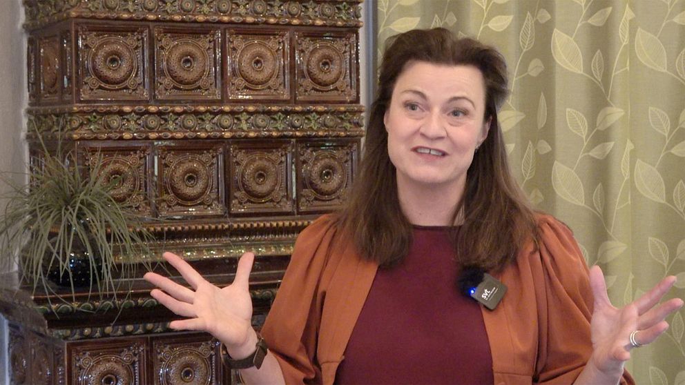 Föreläsaren Charlotte Lindmark gestikulerar med händerna medan hon står framför en inredd bakgrund med ett detaljerat skåp och en växt.Hon bär en orange blus över en röd tröja och har en mikrofon fäst på kläderna. En grön gardin med mönster syns i bakgrunden, vilket skapar en varm atmosfär.