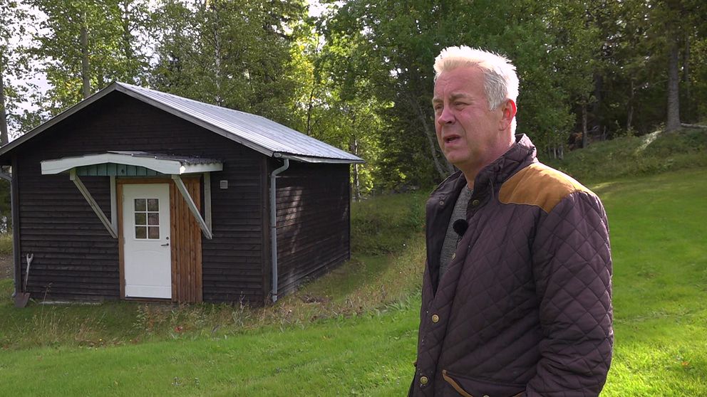 En äldre man i mörklila jacka står framför ett litet hus