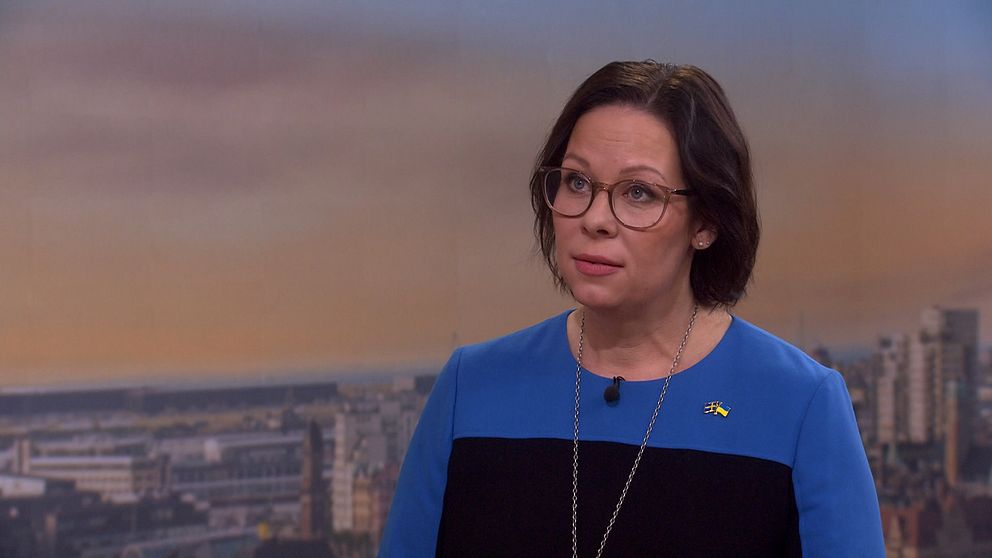 Maria Malmer Stenergard står framträdande i en blå och svart klänning, med ett svenskt flaggmärke synligt. Bakgrunden visar en stadsutsikt med byggnader och en ljus himmel.