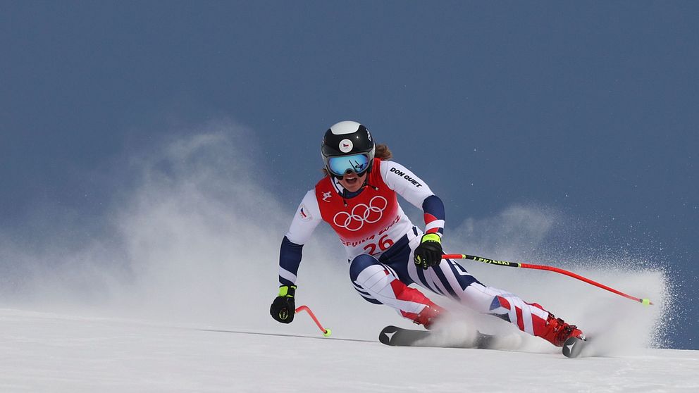 En alpin skidåkare i tävlingsdräkt kör ett åk vid en olympisk skidtävling, med en orange markeringsflagga med olympiska ringar i bakgrunden mot en klarblå himmel. Skidåkaren lutar sig djupt in i en sväng, vilket skapar ett moln av snö.