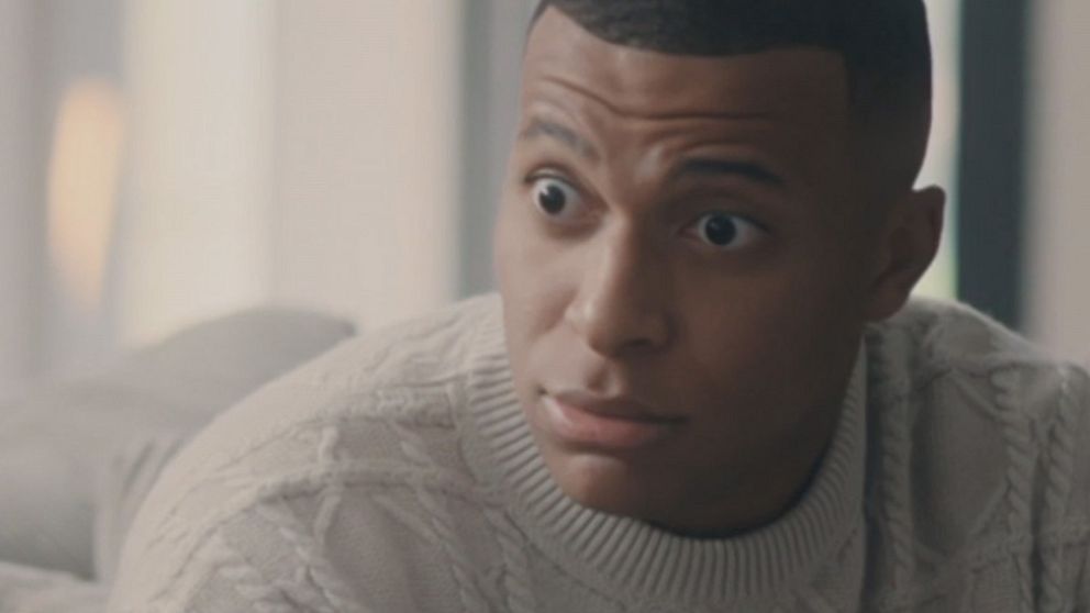 Kylian Mbappé i en vit, stickad tröja sitter på en soffa med en koncentrerad min. Bakgrunden är suddig och ljus, vilket skapar en mjuk och lugn atmosfär.