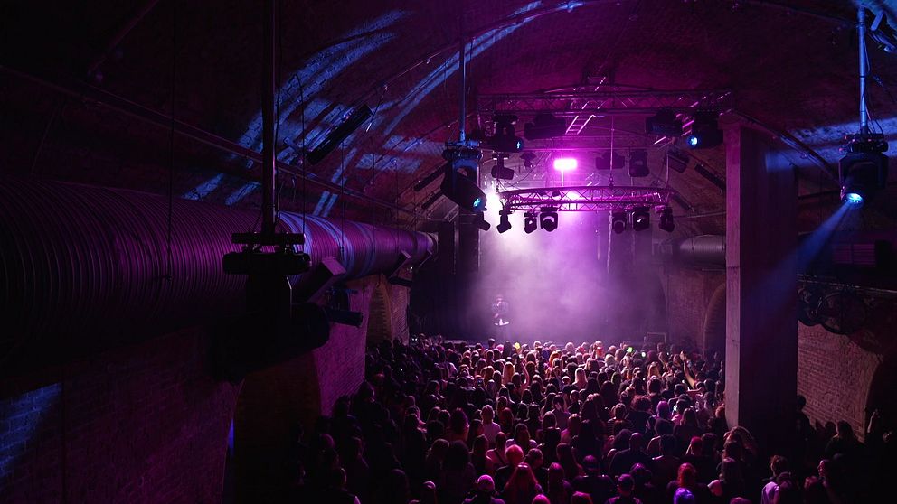 Konsertlokal med tätt packad publik, scenen är upplyst i rosa ljus.