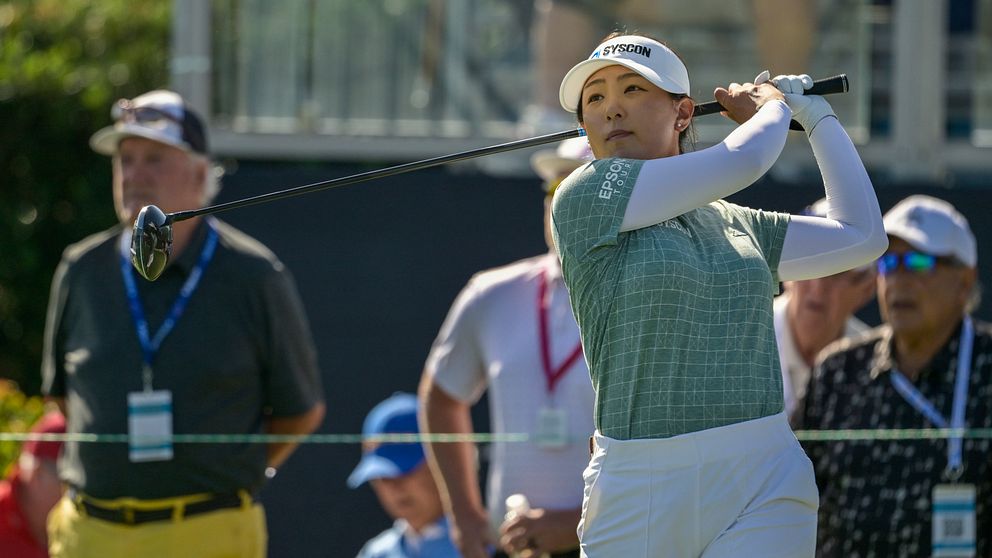 En golfspelare svingar sin driver på en golfbana, omgiven av åskådare som följer spelet. Det är en solig dag, och deltagarna är klädda i lämpliga golfkläder. I bakgrunden syns en del av golfbanan tillsammans med andra personer som är engagerade i spelet.